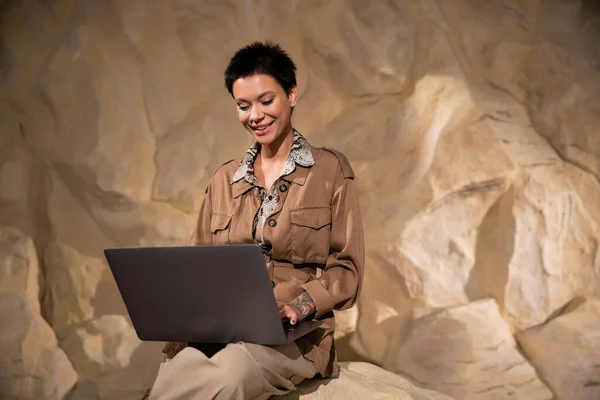 Arqueólogo feliz con tatuaje y pelo corto usando el ordenador portátil en la cueva durante la expedición - foto de stock
