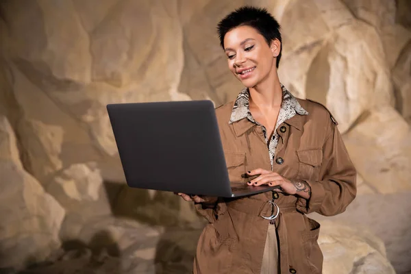 Arqueólogo alegre com tatuagem e cabelo curto usando laptop na caverna durante a expedição — Fotografia de Stock