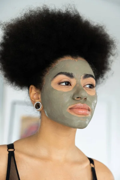 Curly african american woman with clay mask on face looking away at home — Stock Photo