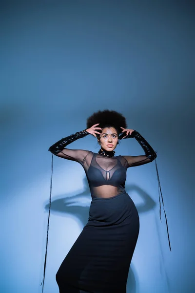 Elegante donna afroamericana in camicia trasparente e reggiseno nero guardando in alto sul blu — Foto stock