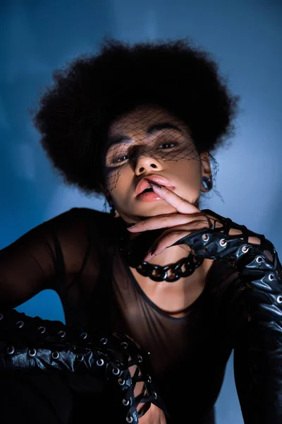 Portrait de femme afro-américaine frisée en voile noir regardant la caméra tout en touchant la lèvre sur le bleu — Photo de stock