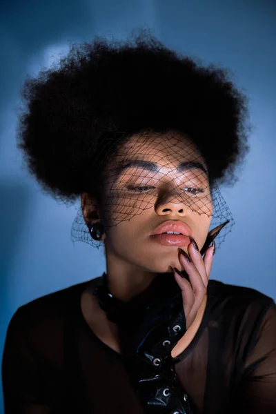 Retrato de mulher afro-americana encaracolado em véu preto olhando para longe ao tocar o rosto no azul — Fotografia de Stock