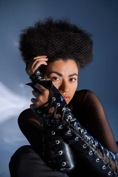 Stylish african american woman in lace-up leather sleeves looking at camera on blue — Stock Photo