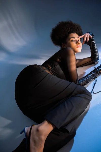 Elegante donna afroamericana in camicia trasparente e maniche in pelle stringata sdraiata sul blu — Foto stock
