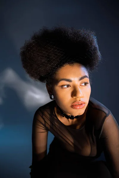 Mujer afroamericana de moda en camisa transparente mirando hacia otro lado en azul - foto de stock