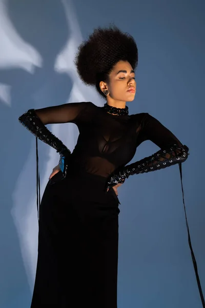 Modelo afroamericano de moda en traje negro posando con las manos en las caderas en azul - foto de stock