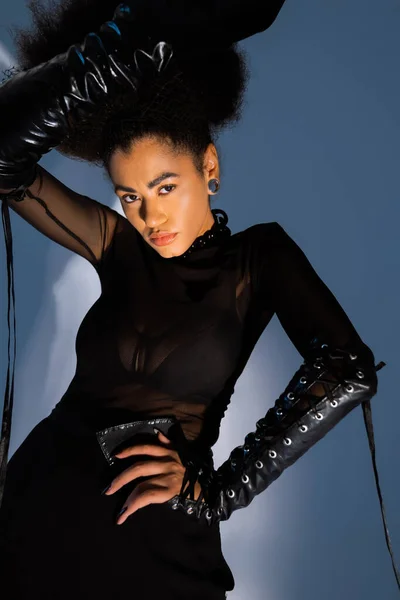 Modèle afro-américain à la mode en tenue noire posant à la main sur la hanche sur bleu — Photo de stock