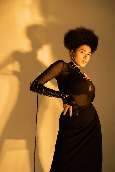 Кучерява афро-американська модель в сексуальній прозорій сорочці і чорний бюстгальтер позує з рукою на стегні на жовтому — стокове фото
