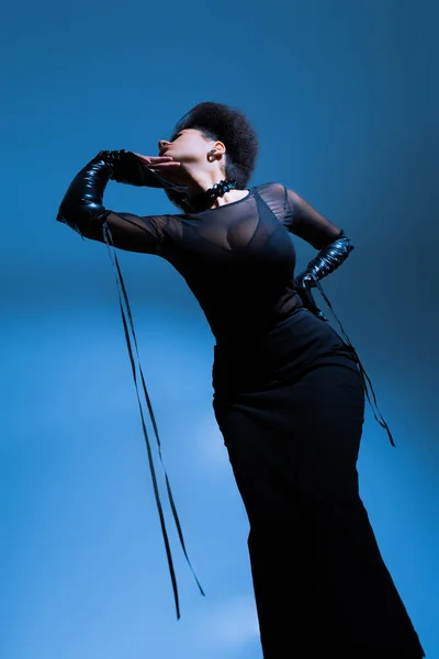 Low angle view of curly african american model in black outfit posing with hand on hip on blue — Stock Photo