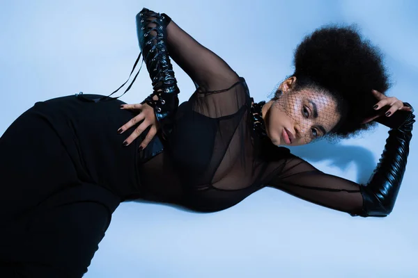 Вид зверху молода афроамериканка в чорній вуаль дивиться на камеру, позує з рукою на стегно на синьому — стокове фото