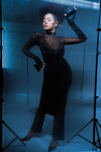 Longitud completa de joven modelo afroamericano en traje negro sexy posando detrás de vidrio con volantes - foto de stock