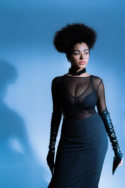 Mujer afroamericana en traje sexy y velo negro mirando hacia otro lado mientras posan en azul - foto de stock