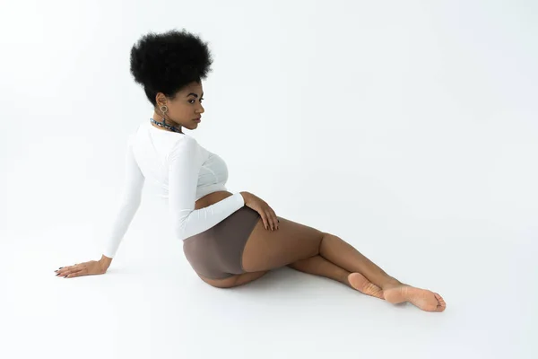 Full length of barefoot african american woman posing in long sleeve shirt and panties while sitting on white — Stock Photo