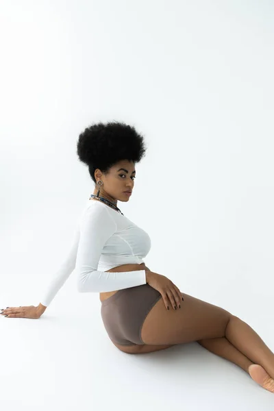 Young african american woman posing in long sleeve shirt and panties while sitting on white — Stock Photo