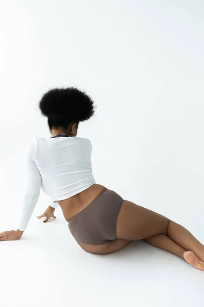 Back view of young african american woman in long sleeve shirt and panties sitting on white — Stock Photo