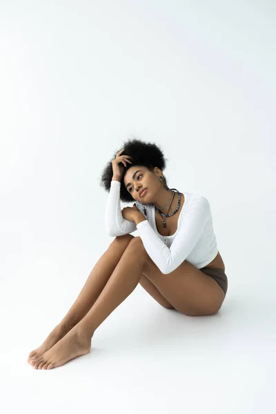 Longitud completa de la soñadora mujer afroamericana en camisa de manga larga y bragas sentado sobre fondo blanco - foto de stock