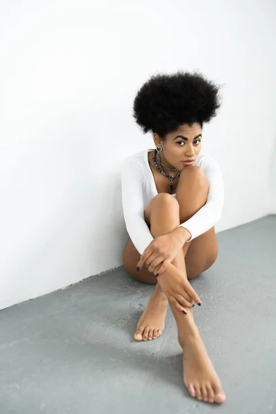 Tutta la lunghezza della donna afroamericana in camicia a maniche lunghe seduta sul pavimento grigio vicino alla parete bianca — Foto stock