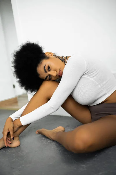 Donna afroamericana a piedi nudi in camicia a maniche lunghe e mutandine seduto sul pavimento grigio vicino alla parete bianca — Foto stock