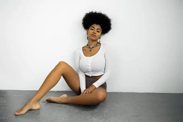 Full length of barefoot african american woman in long sleeve shirt and panties sitting on grey floor near white wall — Stock Photo