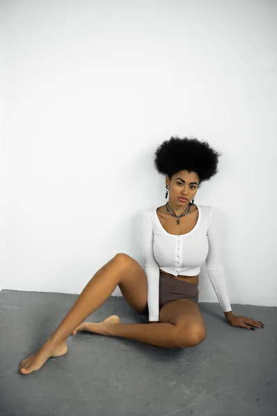 Longitud completa de la mujer afroamericana descalza en camisa de manga larga y ropa interior sentado en el piso gris cerca de la pared blanca — Stock Photo