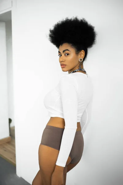 Curly african american woman in long sleeve shirt and underwear standing near white wall — Stock Photo
