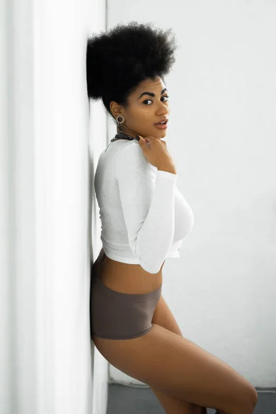 Pretty african american woman in long sleeve shirt and underwear standing near white wall — Stock Photo
