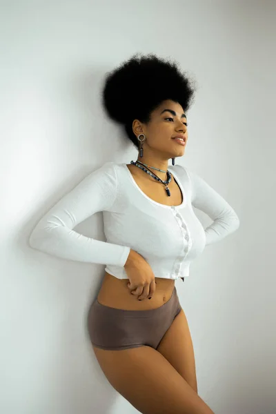 Cheerful african american woman in long sleeve shirt and underwear standing near white wall — Stock Photo