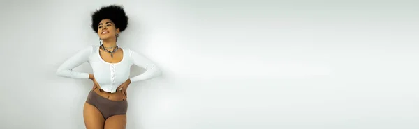 Heureuse femme afro-américaine en chemise à manches longues et sous-vêtements debout avec les mains sur les hanches près du mur blanc, bannière — Photo de stock