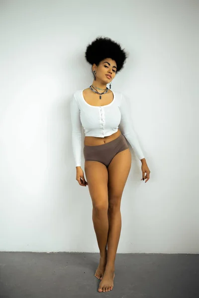Piena lunghezza della donna afroamericana a piedi nudi in camicia a maniche lunghe e mutandine in piedi vicino alla parete bianca — Foto stock