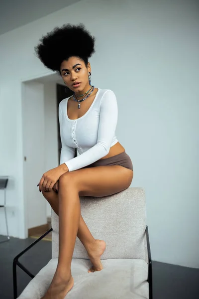 Young african american woman in long sleeve shirt and panties looking away while sitting on armchair near white wall — Stock Photo