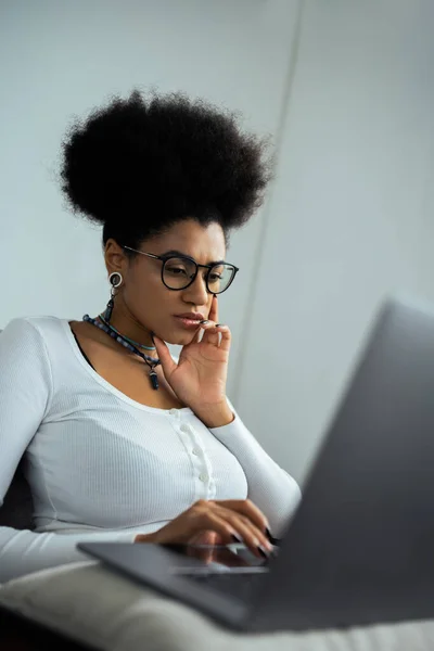 Femme afro-américaine concentrée dans des lunettes en utilisant un ordinateur portable à la maison — Photo de stock