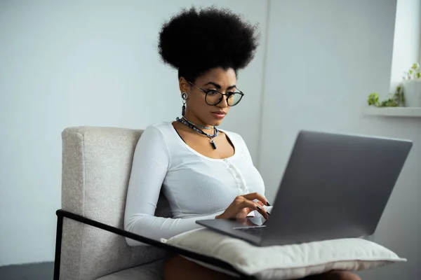 Mulher americana africana concentrada em óculos usando laptop enquanto sentado em poltrona — Fotografia de Stock