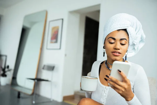 Mujer afroamericana bonita con toalla en la cabeza usando un teléfono inteligente y sosteniendo una taza de café - foto de stock