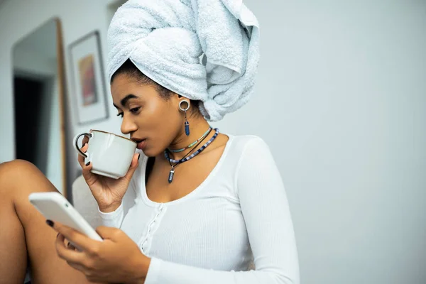 Mujer afroamericana bonita con toalla en la cabeza usando teléfono inteligente y beber café - foto de stock