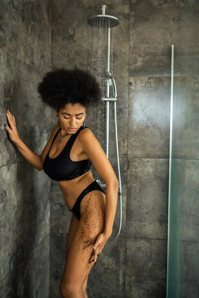Curly african american woman applying coffee scrub under water in shower cabin — Stock Photo