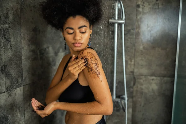 Rizado africano americano mujer en crop top masaje hombro con café exfoliante en ducha - foto de stock