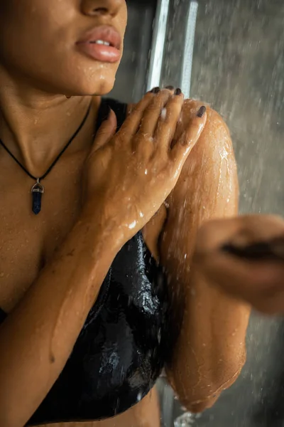 Cropped view of african american woman holding blurred scrub while standing under water in shower — Stock Photo