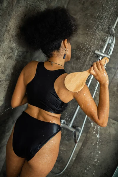 Вид ззаду кучерявої афроамериканської жінки, що масажує тіло пензлем у душі — стокове фото