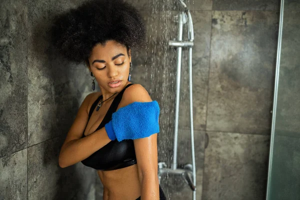 Кучерява афроамериканка масажує тіло рукавичкою в душовій кабіні — стокове фото