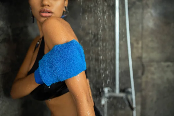 Vista recortada de mujer afroamericana masajeando brazo con guante mientras toma ducha - foto de stock
