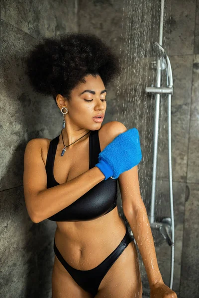 Jeune femme afro-américaine nettoyage du corps avec un gant de massage dans la douche — Photo de stock