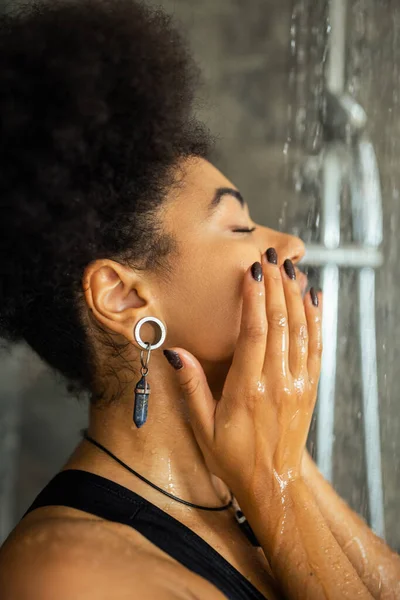 Vista lateral da mulher afro-americana lavando o rosto debaixo d 'água no chuveiro — Fotografia de Stock