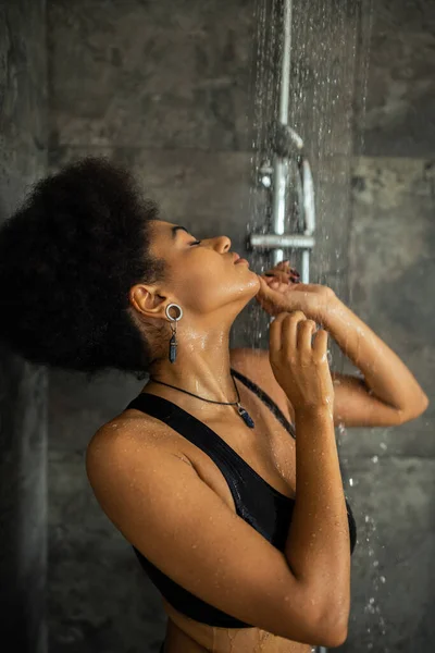 Vista laterale della giovane donna afroamericana in piedi sotto l'acqua in cabina doccia — Foto stock