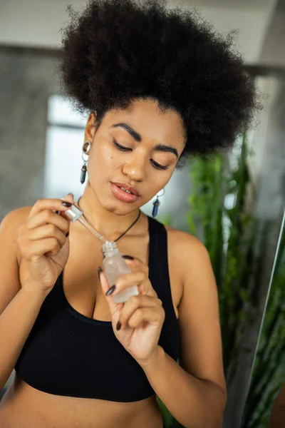Curly african american woman in crop top holding blurred serum in bathroom — Stock Photo