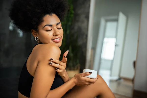 Mulher americana africana agradável aplicando creme cosmético no ombro no banheiro — Fotografia de Stock