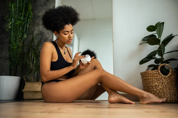 Joven mujer afroamericana sosteniendo crema cosmética mientras está sentada en el suelo en el baño - foto de stock