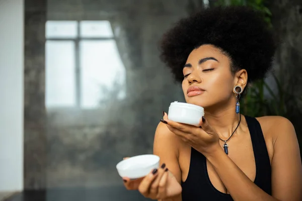 Mujer afroamericana joven oliendo crema cosmética en el baño - foto de stock