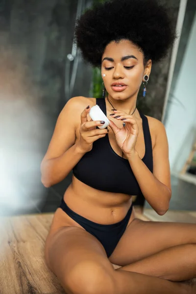 Jeune femme afro-américaine avec crème cosmétique sur la joue tenant le récipient dans la salle de bain — Photo de stock