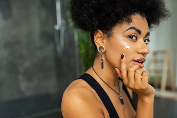 Femme afro-américaine appliquant crème sur le visage dans la salle de bain floue — Photo de stock