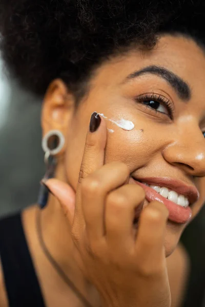 Vista recortada de la sonriente mujer afroamericana aplicando crema en la mejilla en casa - foto de stock